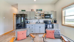 Cocina compacta con armarios grises, microondas y cafetera. Hay dos sillones grises con cojines naranja y un banco de madera.