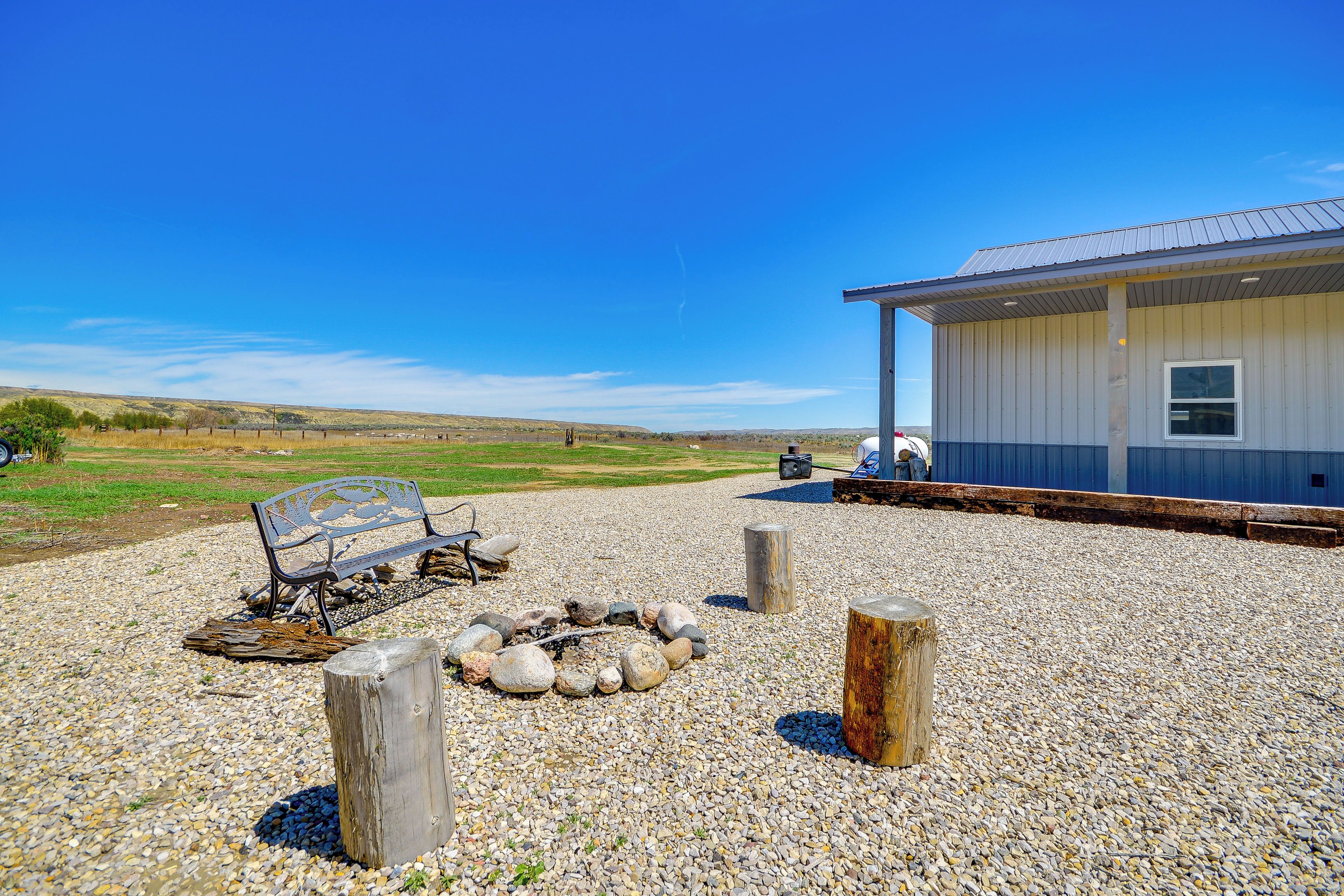 A building with a metal roof and a window, surrounded by a gravel area with a fire pit and a bench.
