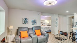 Een moderne woonkamer met een grijze leren bank, twee oranje kussens, een kleine ronde tafel met een lamp, en een houten eettafel met twee stoelen.