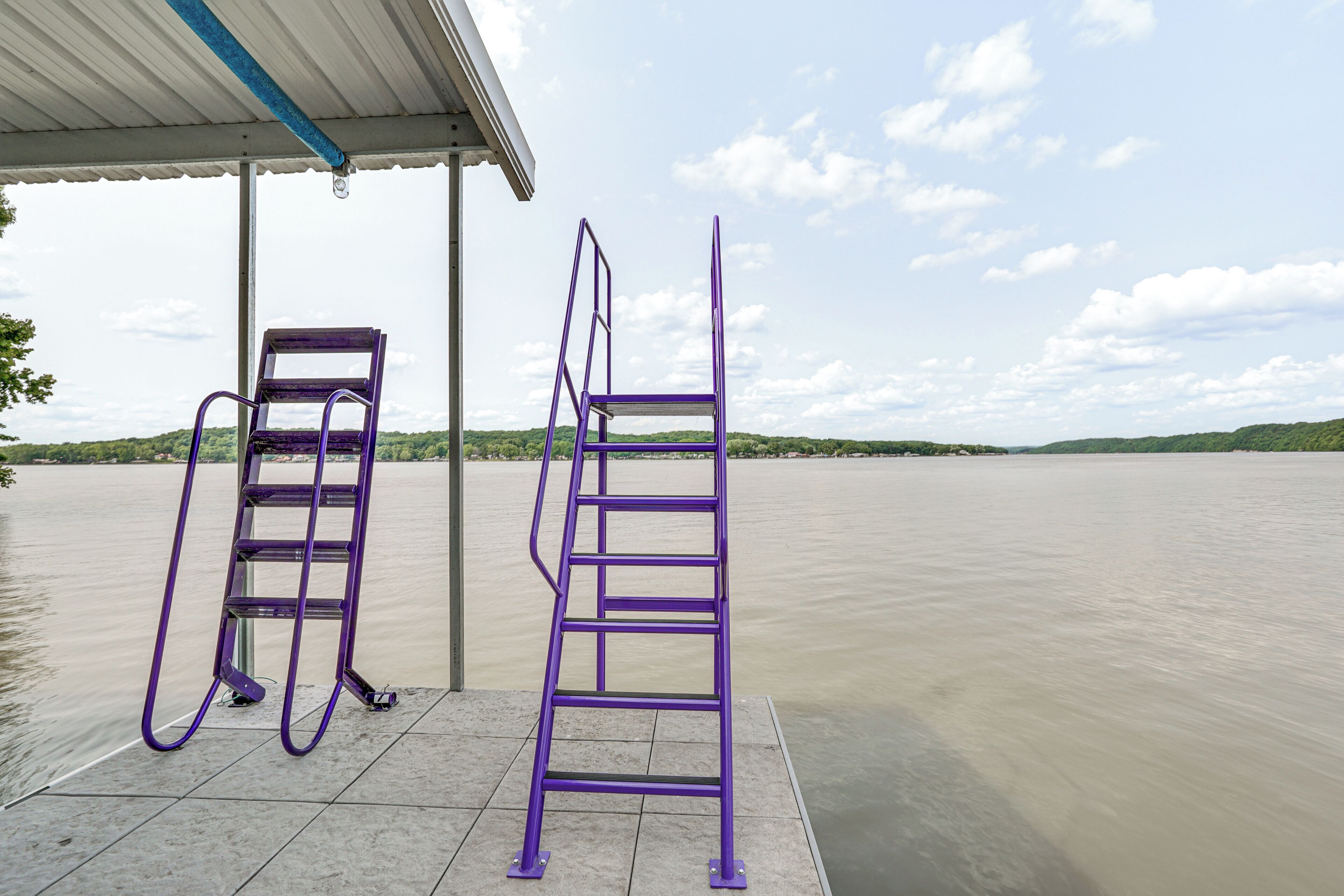 Due scalette metalliche viola su un molo, con un'acqua calma e un paesaggio verde in lontananza.