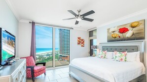 A hotel room with a bed, a TV, a chair, a ceiling fan, and a large window with a view of buildings and a beach. - Tower 3-0904 Stunning Sunset Views! (Pensacola Beach)