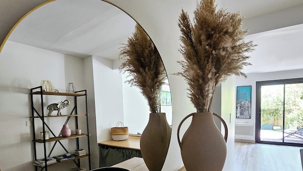 A modern interior with a wooden table, two large vases with dried plants, a round mirror, and a bookshelf with various items. - Jardin Privé, Parking Free, Centre Cassis (Cassis)