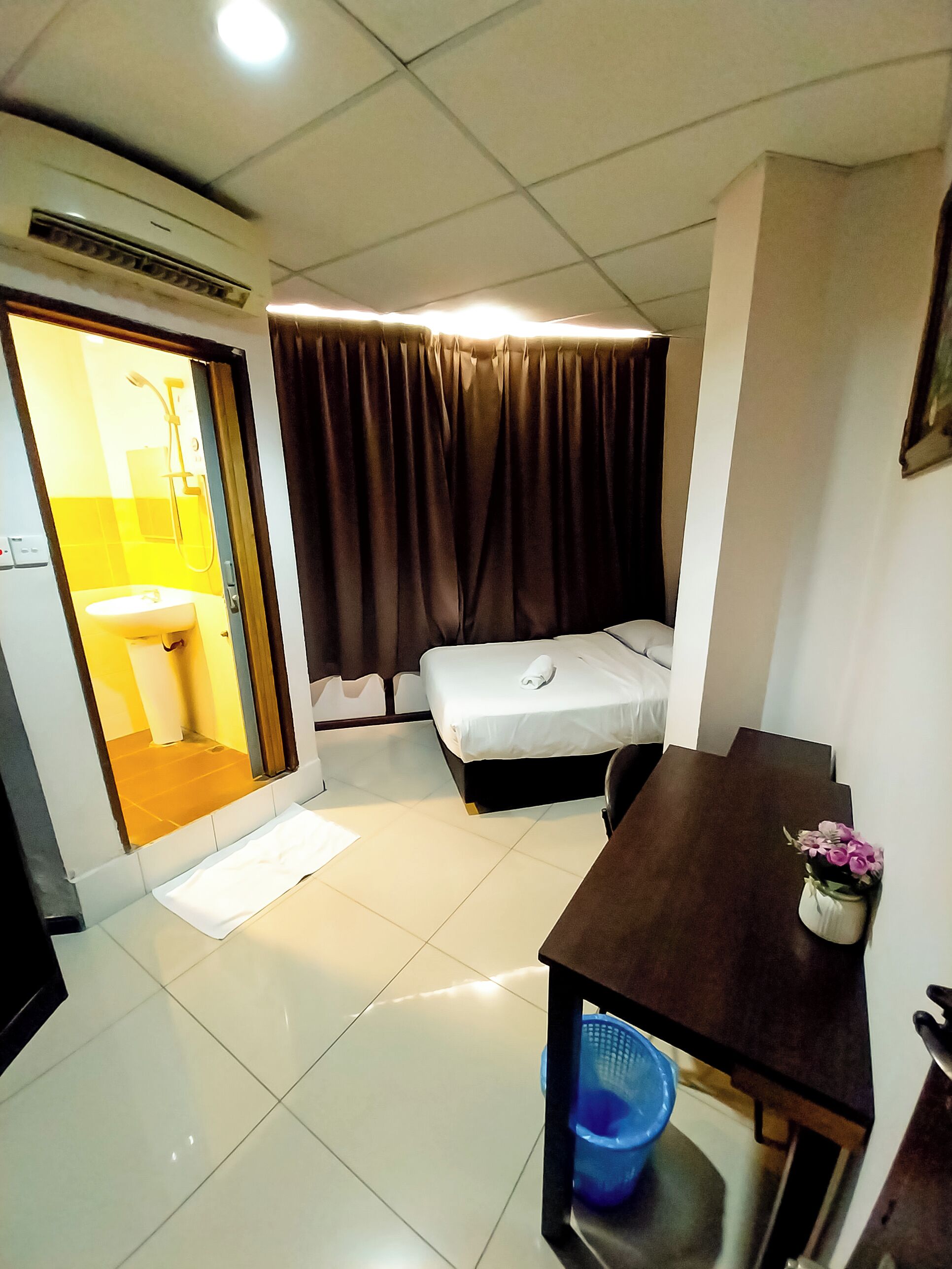 Une chambre d’hôtel de taille modeste, comprenant un lit, une petite table, une poubelle bleue et une salle de bain visible à travers une porte ouverte.