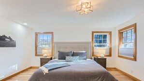 A bedroom with a large bed, two bedside tables, a modern chandelier, and large windows with blinds.