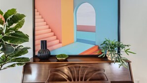A framed abstract painting with geometric shapes and a staircase design, placed on a wooden desk with a vase and a small plant.