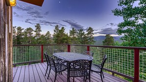 A wooden deck with a round table and chairs, surrounded by a red railing, overlooking a forested landscape.