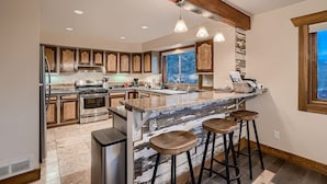 A modern kitchen with wooden cabinets, a large island with stools, stainless steel appliances, and a large window providing a view.