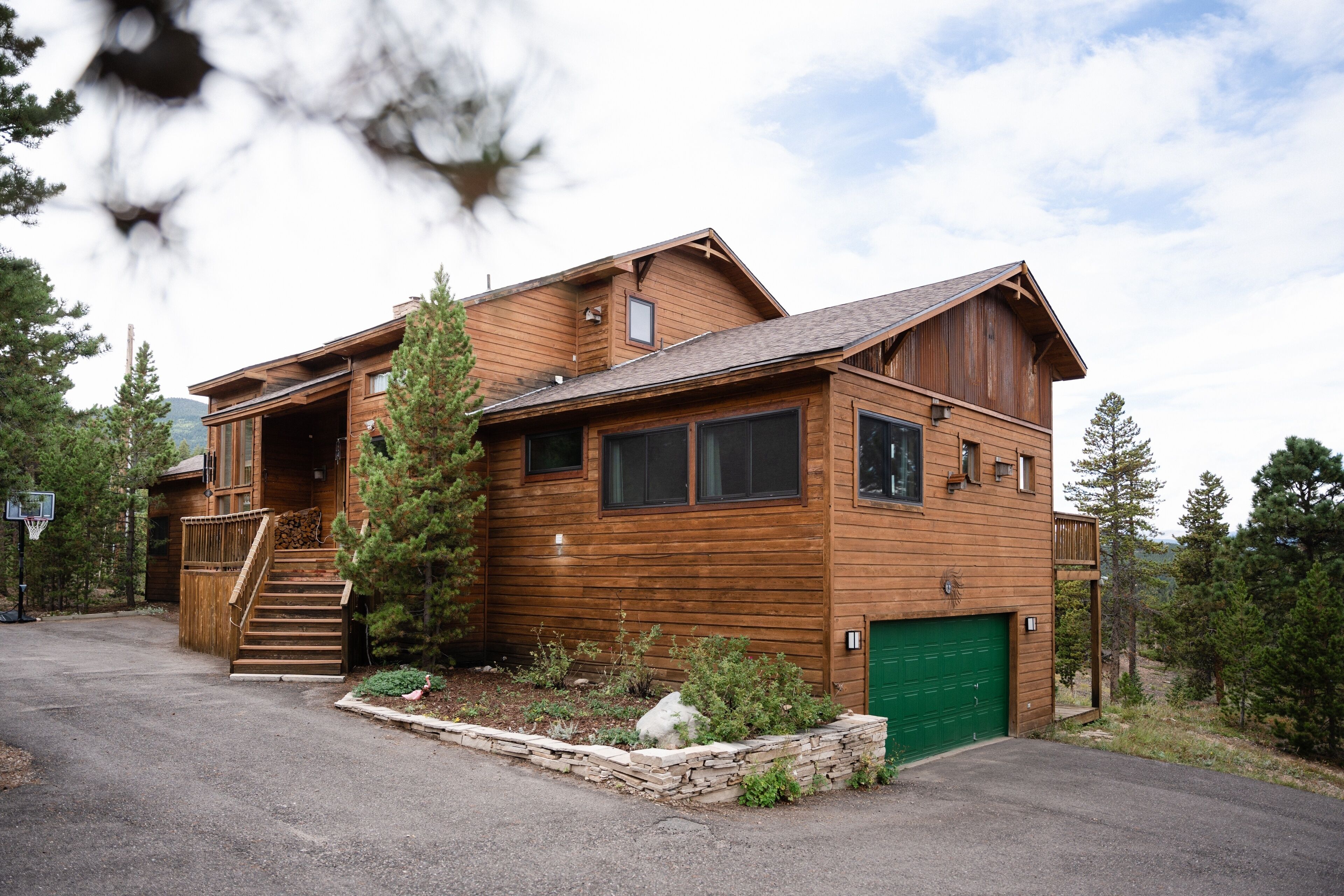 A two-story wooden cabin with a green garage, a staircase leading to the upper level, and a small garden area with rocks and plants.