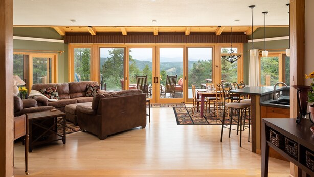 A spacious living area with a large sofa, dining table, and chairs, a kitchen area with a sink and counter, and large sliding glass doors leading to a scenic outdoor view. - Lookout Lodge In Wondervu 4 Bedroom Home by RedAwning (Golden)
