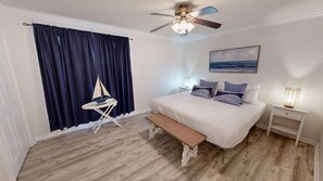 Un dormitorio con una cama, un banco, un modelo de velero, un ventilador de techo, una mesita de noche con una lámpara y un cuadro en la pared.