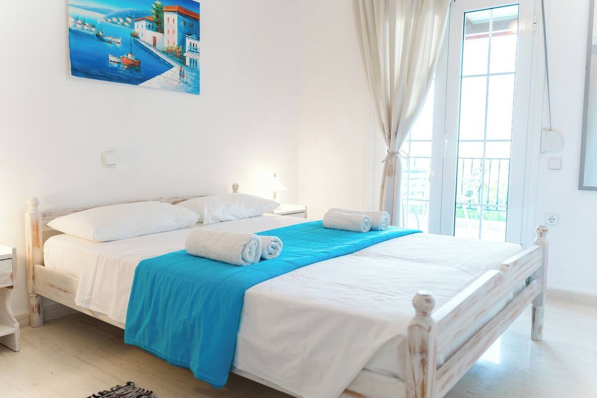 A neatly made bed with white and blue bedding, a painting of a coastal scene on the wall, and a window with curtains.