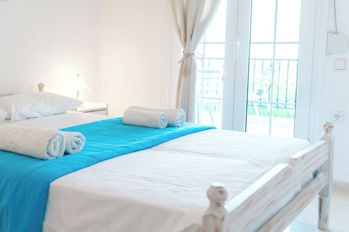 A neatly made bed with white linens and a blue bedspread, a bedside table with a lamp, and a view of the outdoors through a large window.