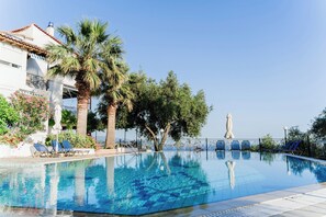 A clear swimming pool with lounge chairs, palm trees, and a building with a tiled roof. - Villa Kallithea (Parga)