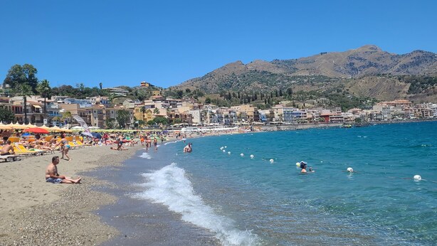 A beach with clear blue water, pebbly shore, and a mountainous backdrop. - The balcony overlooking the sea + parking space
 (Giardini Naxos)