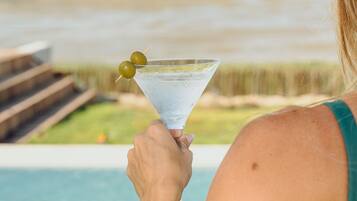 Uma pessoa segurando uma taça de martini com líquido transparente e duas azeitonas, ao lado de uma piscina com vista para um corpo d'água e vegetação ao fundo.