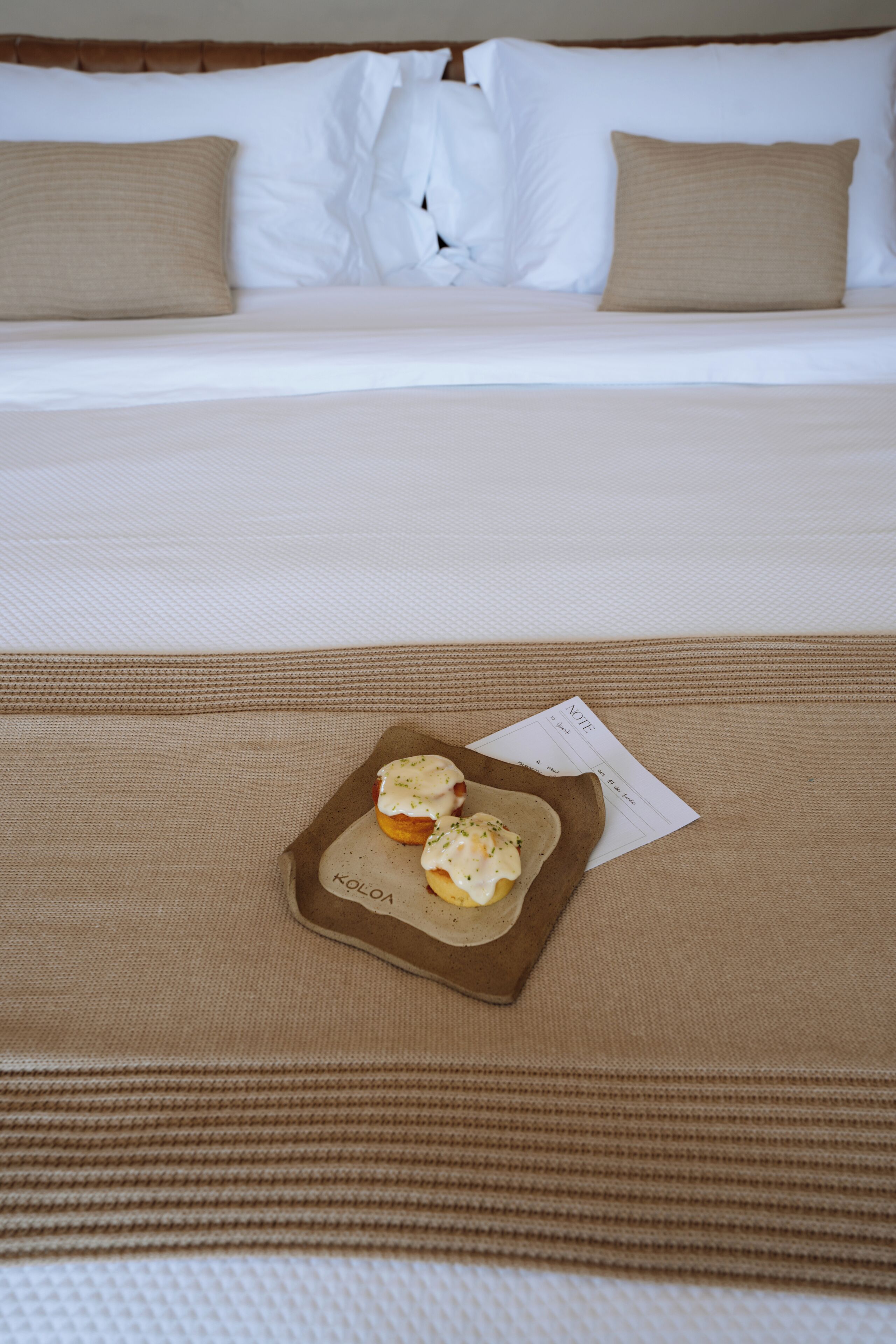 A neatly made bed with a brown bedspread and two pillows, a tray with two pastries, and a note on the bed.