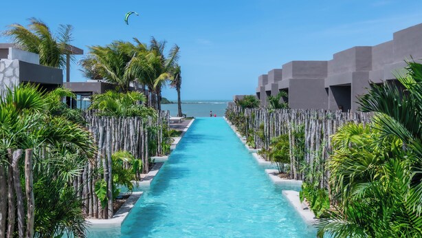 Piscina azul clara com um caminho reto que leva ao mar, rodeada por palmeiras e edifícios modernos.