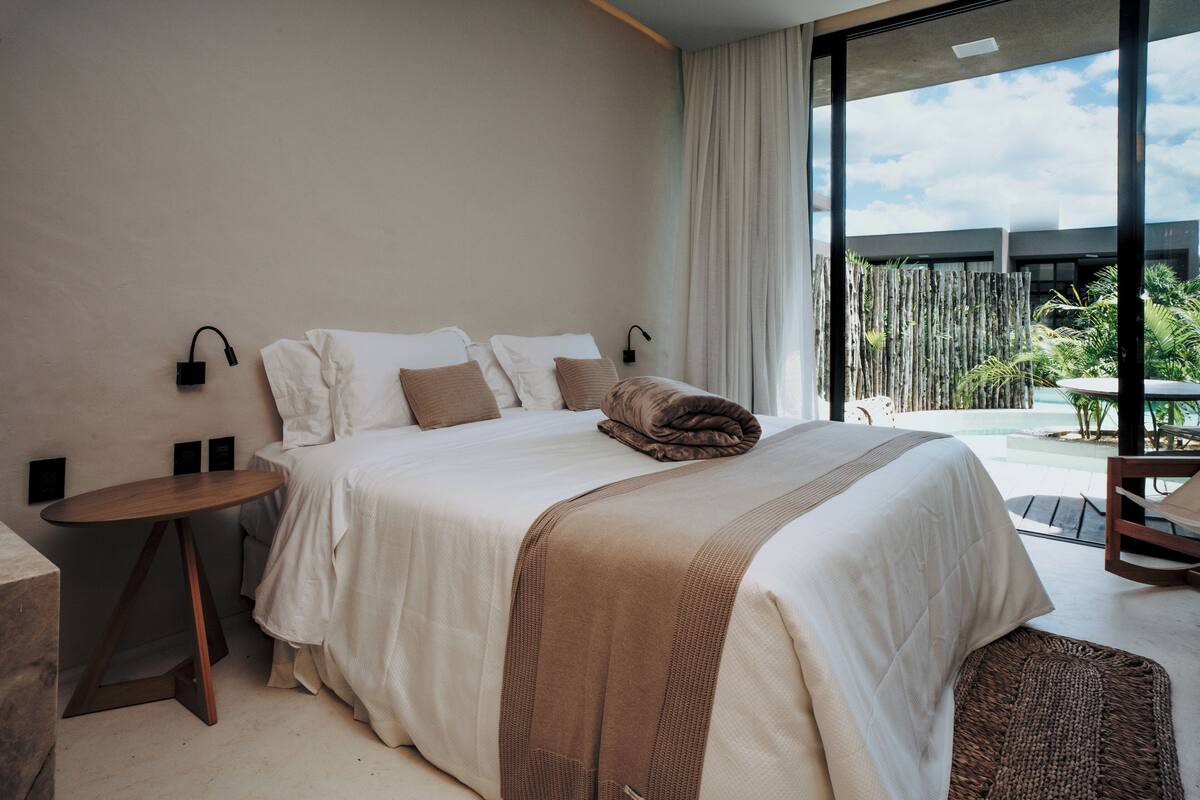 A modern bedroom with a large bed, a bedside table, and a view of an outdoor pool area.