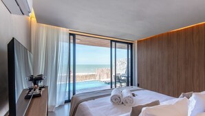Quarto de hotel com uma cama grande, televisão, escrivaninha de madeira e vista para a praia e oceano.