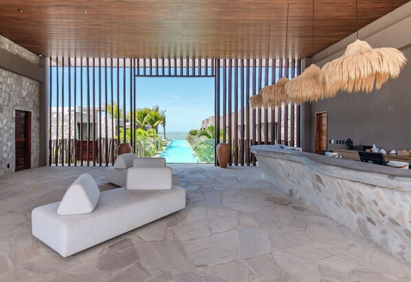 Un interior moderno con suelo de piedra, un sofá blanco, un mostrador de recepción y vistas a una piscina y palmeras.