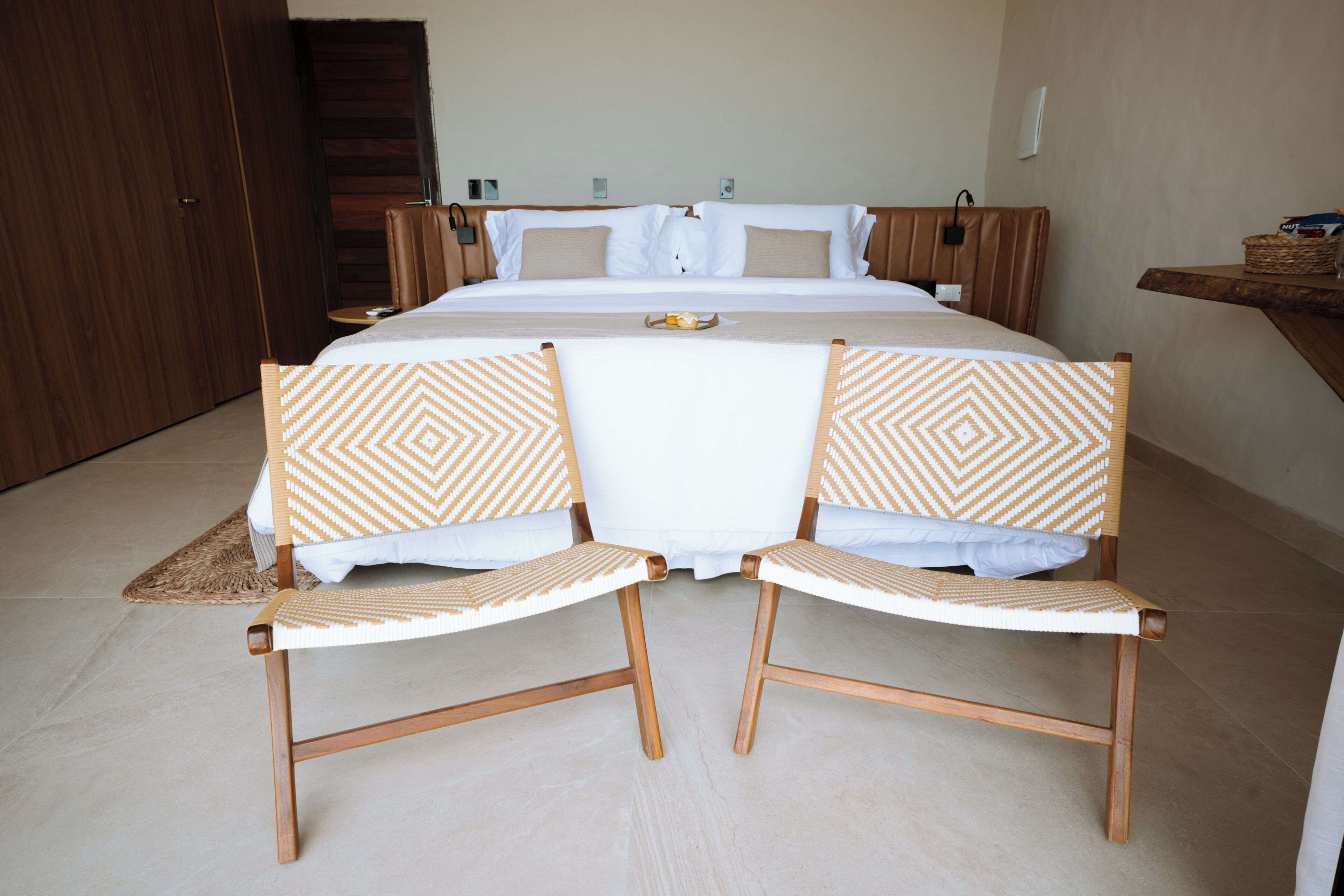A bedroom with a large bed, two upholstered chairs with patterned cushions, a wooden headboard, and a bedside table with a basket.