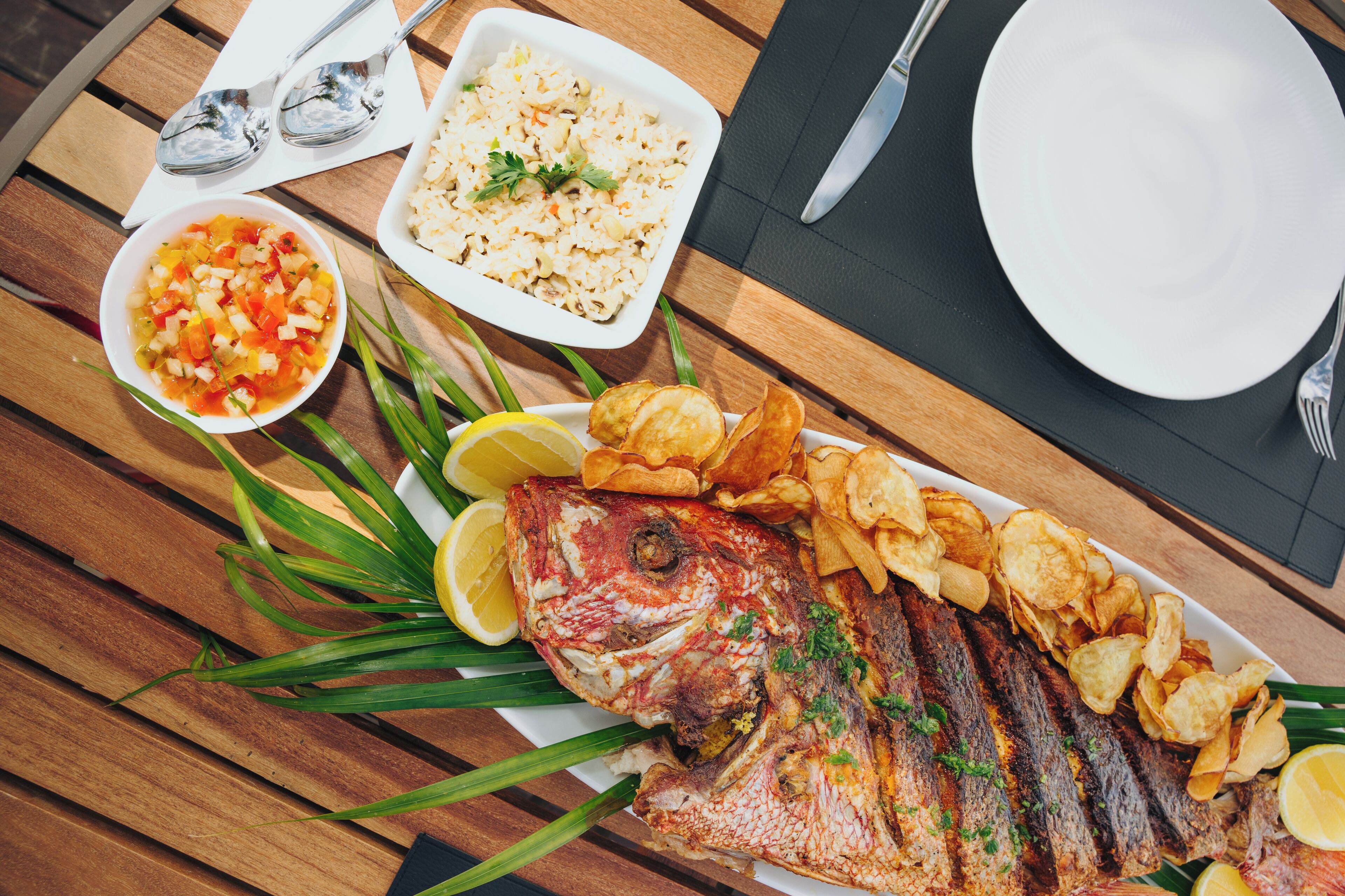 A wooden table with a whole grilled fish, lemon slices, and a side of chips.