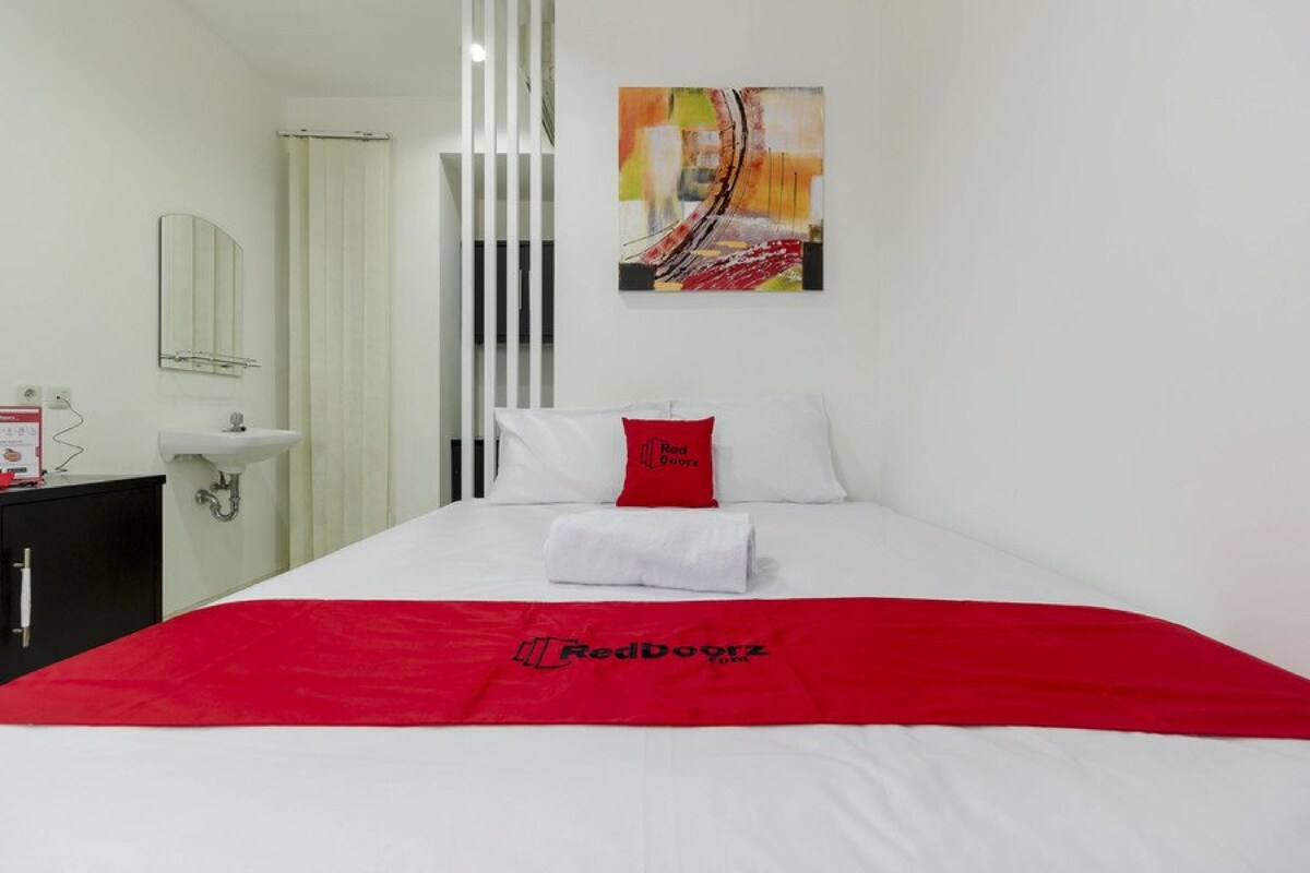 A hotel room with a bed, a red blanket, a white pillow, a red pillow with 'Red Doors' text, a white wall, a painting, a bathroom with a sink and mirror, and a black cabinet.