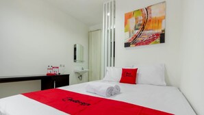A hotel room with a bed, a red and white bedspread, a white pillow with a red logo, a white wall, a painting, a mirror, a sink, and a red object on the counter. - Reddoorz Plus @ Pasar Baru (Jakarta)