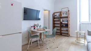 A compact living space with a flat-screen TV mounted on the wall, a wooden bookshelf, a dining table with two chairs, and a small round table with a lamp.