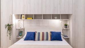 A bedroom with a bed, a headboard with shelves, a bedside table, a lamp, and a hanging plant.