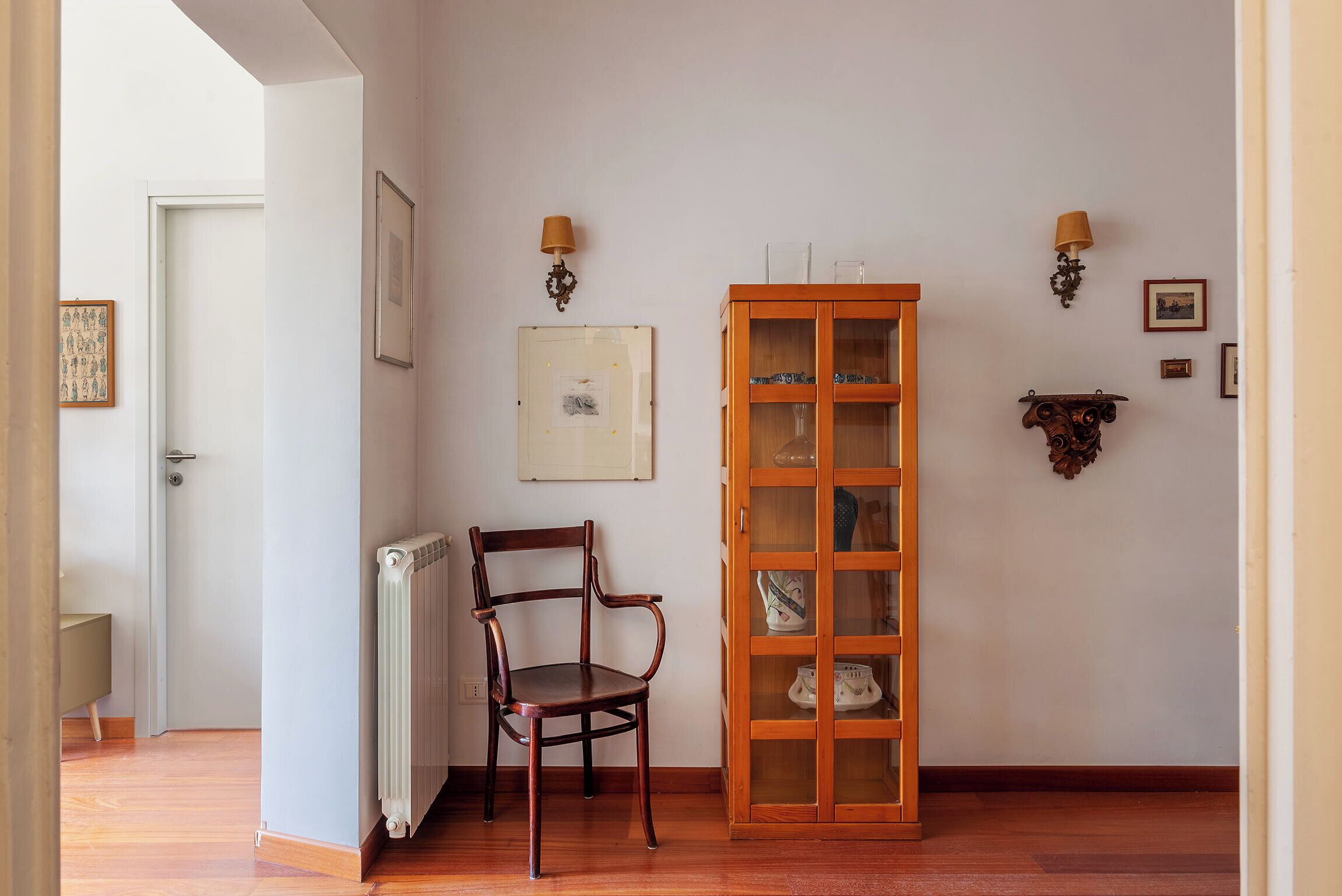 A room with a wooden chair, a cabinet with glass doors, and wall-mounted lamps.