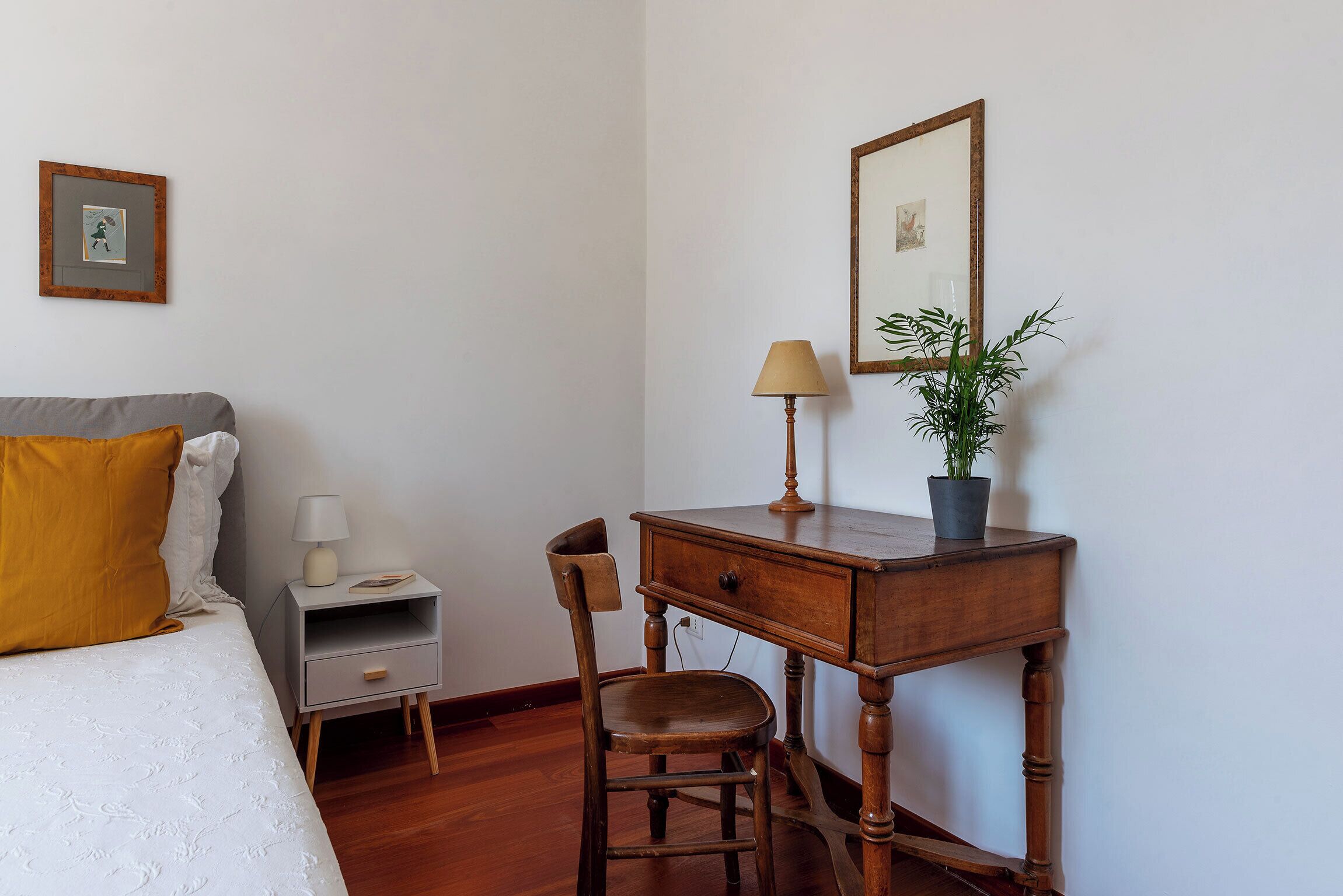 A bedroom with a bed, a nightstand, a desk, a chair, a lamp, and a potted plant.