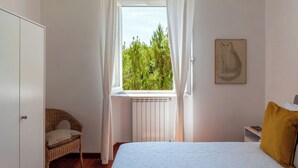 A bedroom with a bed, a wicker chair, a radiator, and a window with curtains.
