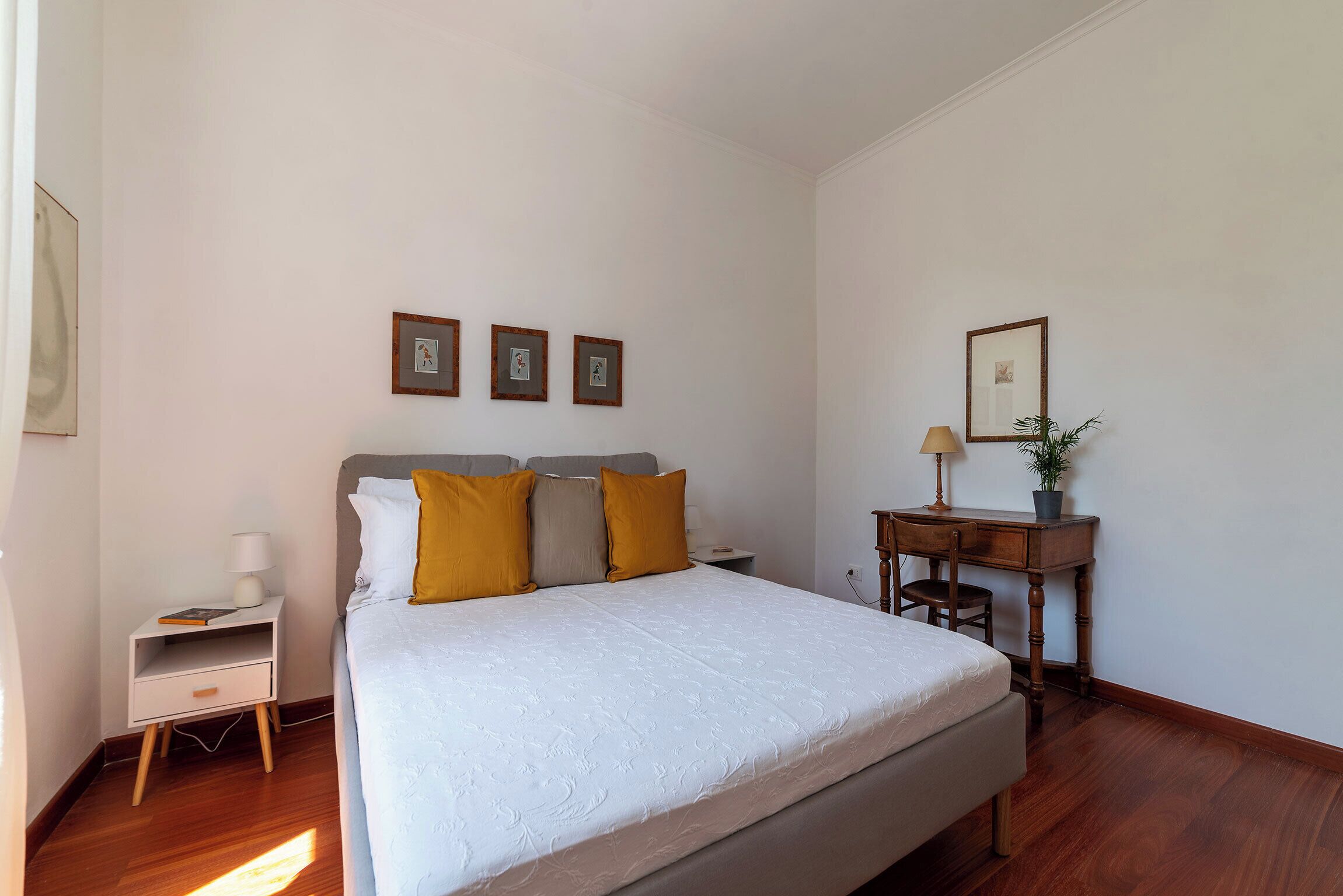 Un dormitorio con una cama grande, una mesita de noche, un escritorio y una silla. Hay tres cuadros enmarcados en la pared.