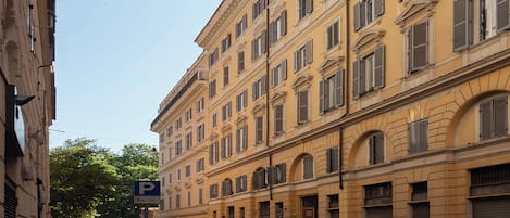 A narrow street lined with parked cars and a row of yellow buildings with multiple windows and shutters.