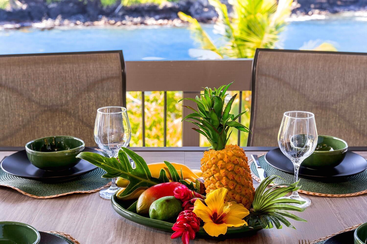 Une table dressée avec un ananas, une mangue et une fleur d’hibiscus, ainsi que deux verres et des bols, le tout posé sur une table en bois donnant sur une plage.