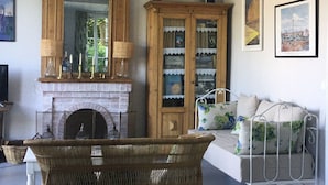 A cozy room with a fireplace, a wicker chair, a sofa with floral cushions, a wooden cabinet with glass doors, and framed pictures on the wall.