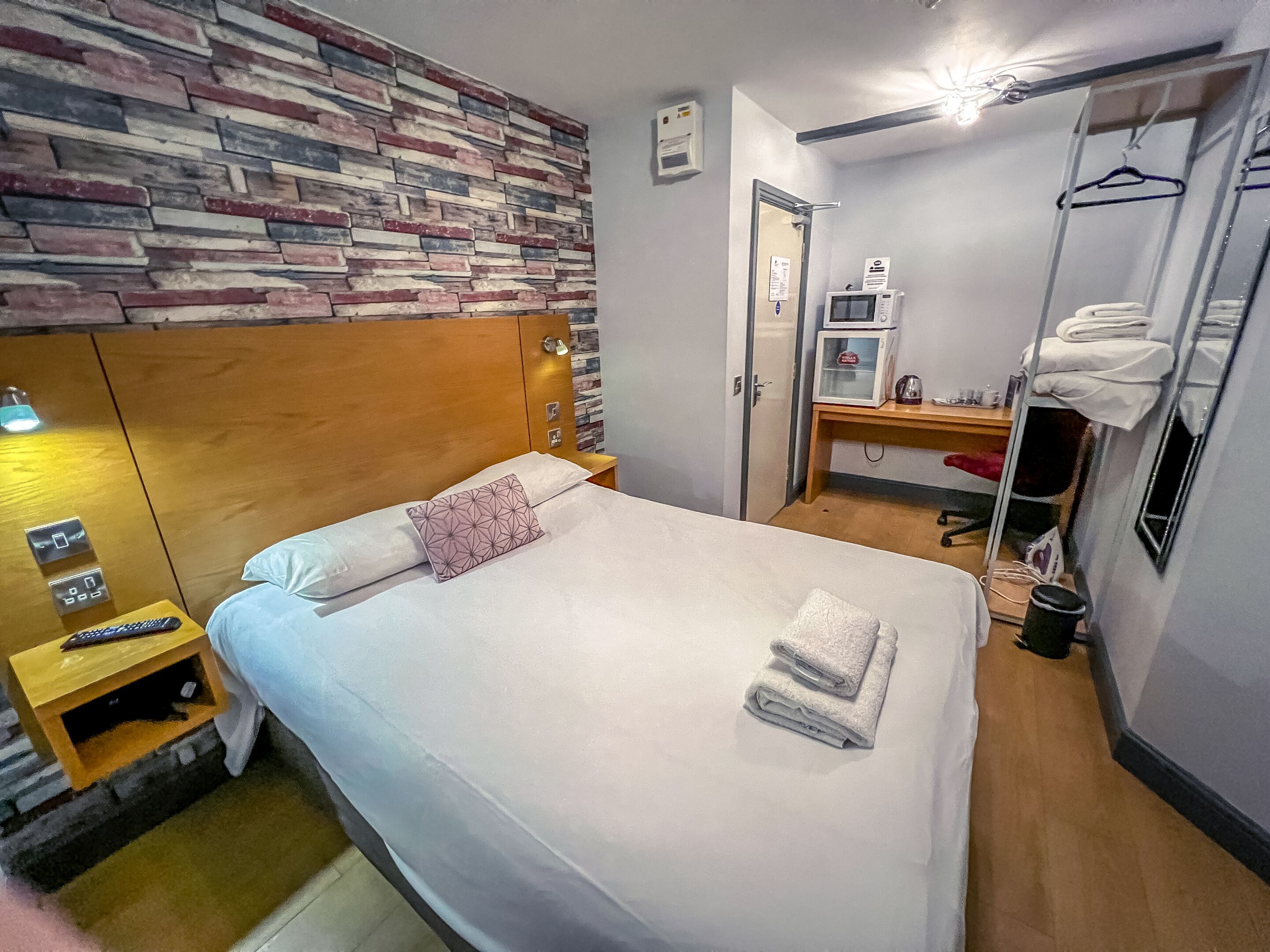 A hotel room with a bed, a wooden headboard, a nightstand with a remote control, a microwave, a small desk, and a wardrobe.
