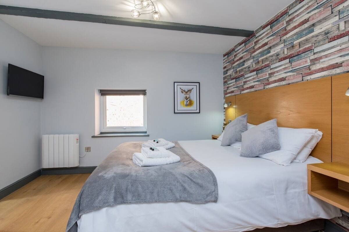 A bedroom with a bed, a window, a radiator, a wall-mounted TV, and a brick-patterned wall.