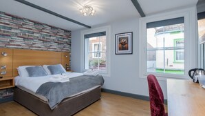 Un dormitorio moderno con cabecera de madera, una cama grande con ropa de cama gris, una silla roja y vistas a un edificio de ladrillo a través de las ventanas.