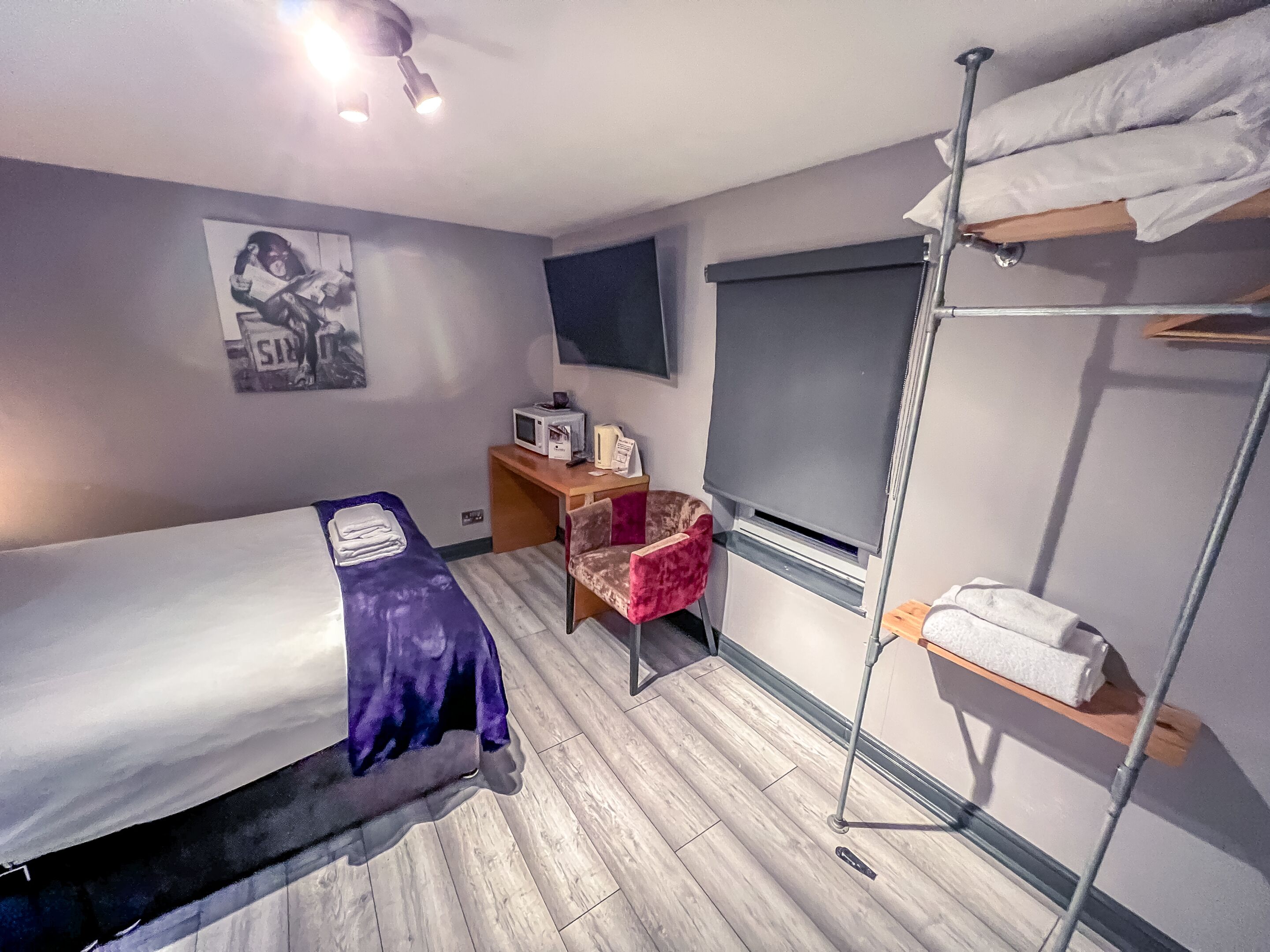 A compact hotel room with a single bed, a desk with a computer, a chair, a TV, and a shelf with towels.