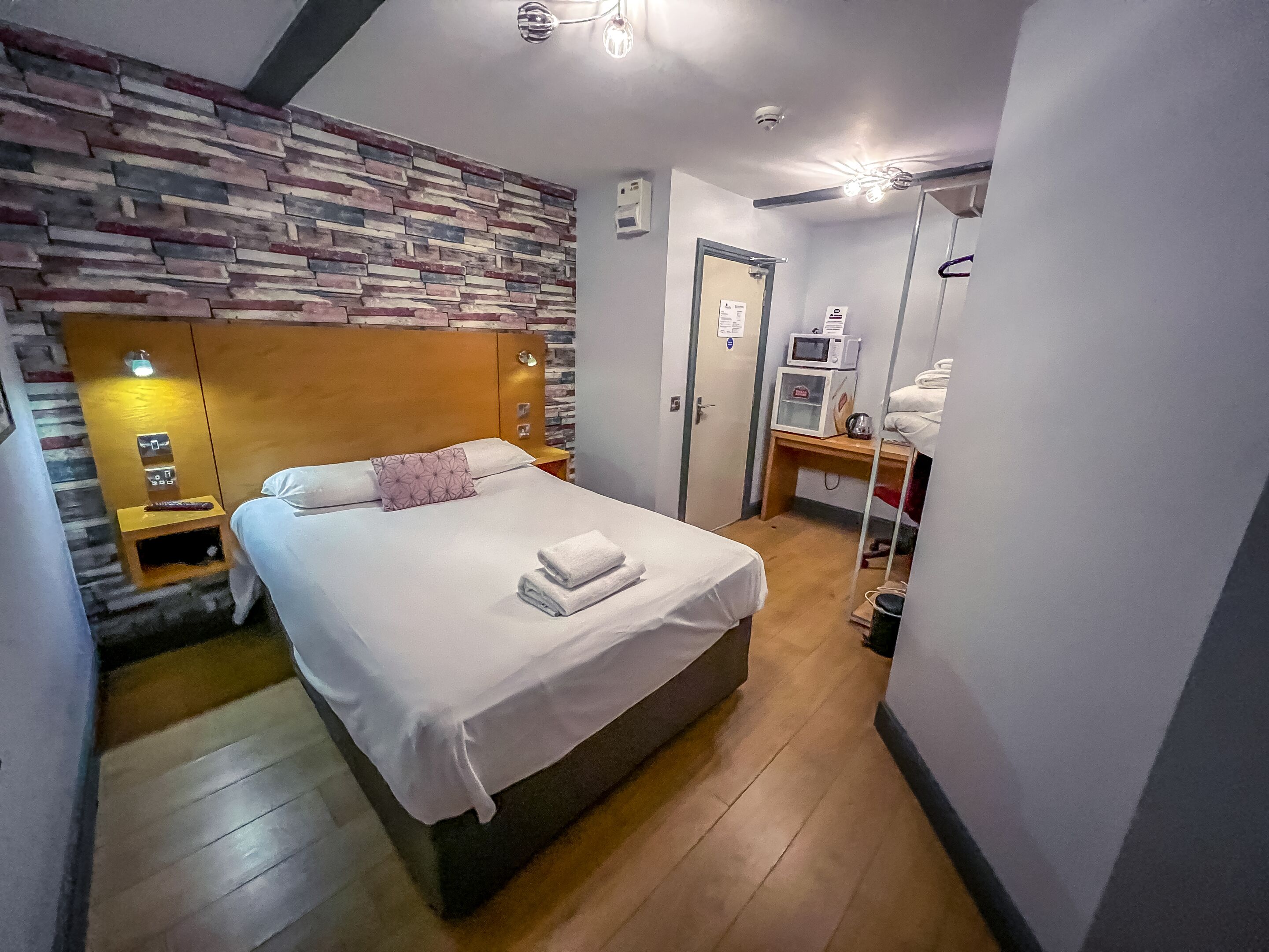 Un dormitorio con cama, cabecero de madera, pared con diseño de piedra, mesita de noche con lámpara y un taburete pequeño de madera.