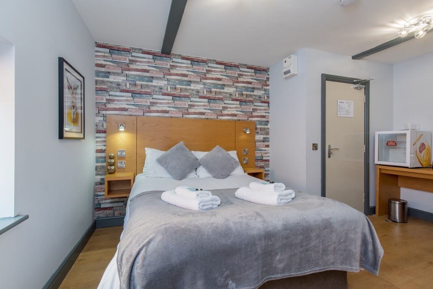 A neatly arranged hotel room with a bed, a wooden headboard, a brick-patterned wall, a framed picture, a bedside table with a lamp, and a microwave on a wooden counter.