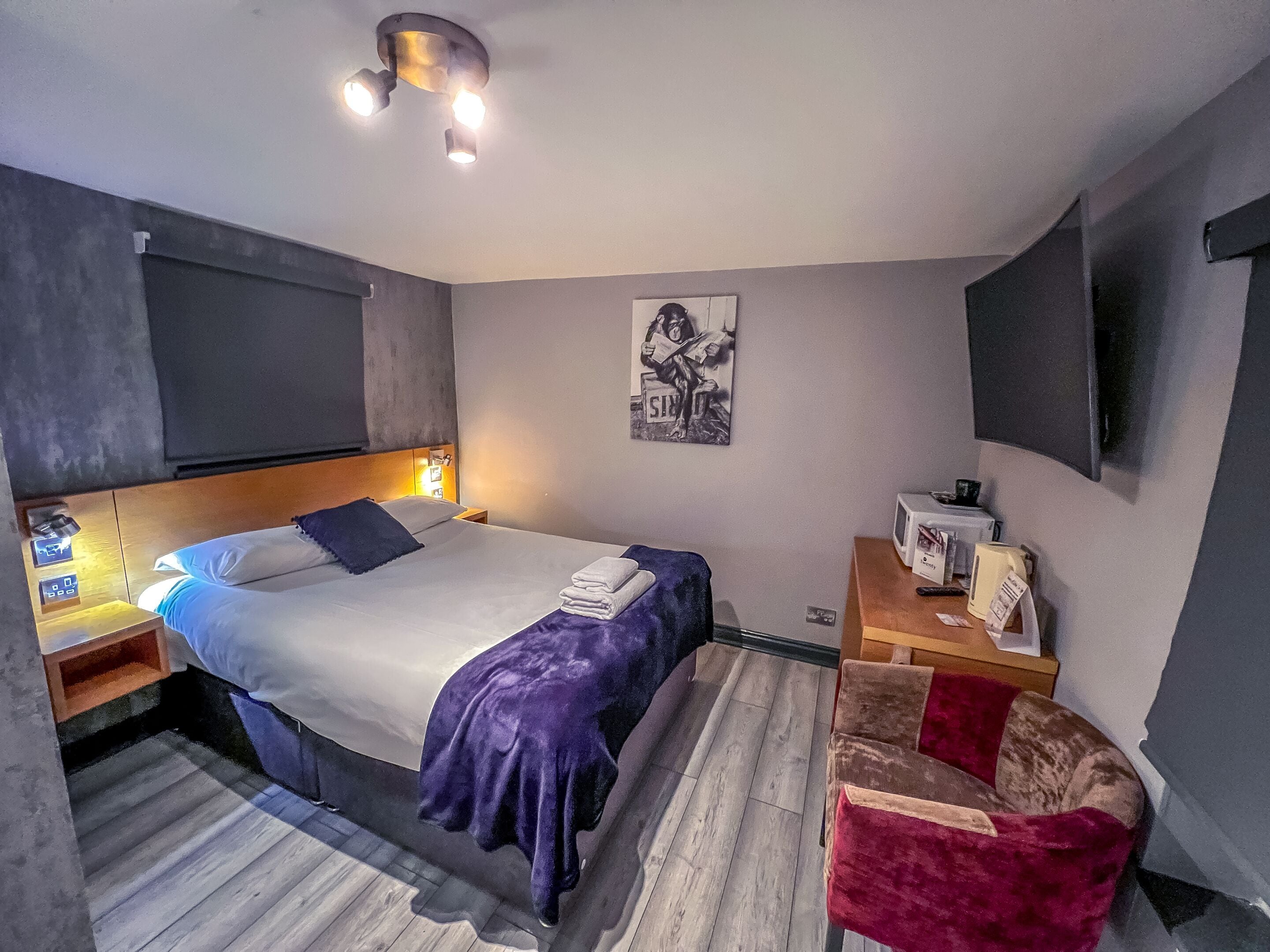 A modern hotel room with a large bed, bedside tables, a small desk, a chair, and a flat-screen TV mounted on the wall.