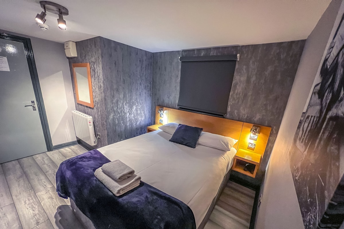 A modern hotel room with a large bed, a wooden headboard, a nightstand with a lamp, a radiator, and a door with a handle.