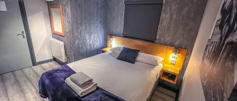 A modern hotel room with a large bed, a wooden headboard, a nightstand with a lamp, a radiator, and a door with a handle.
