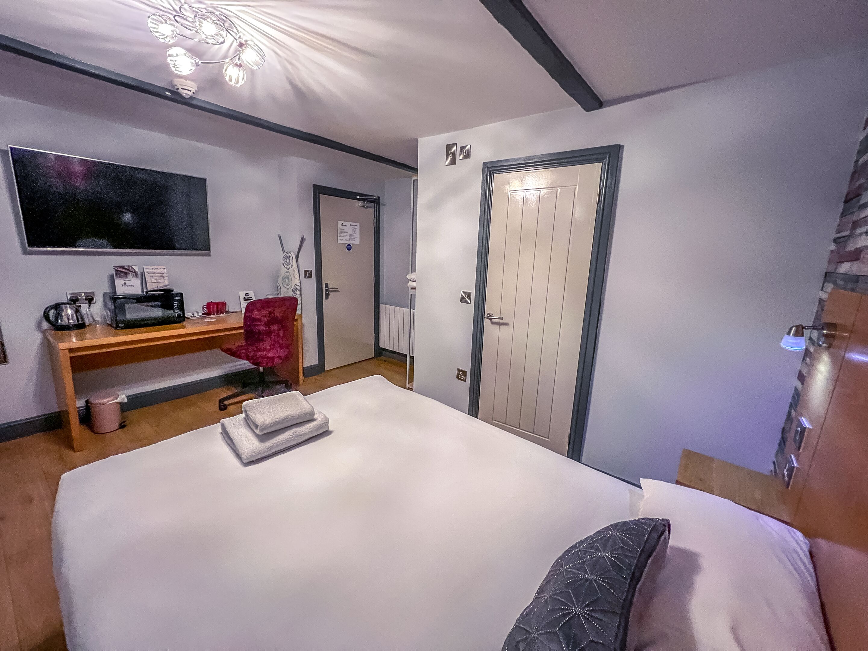 Quarto de hotel com cama, escrivaninha, computador, cadeira, televisão e porta.