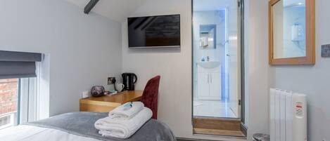 Quarto moderno com cama, televisão na parede, uma cadeira vermelha, penteadeira de madeira e banheiro visível por uma porta aberta.