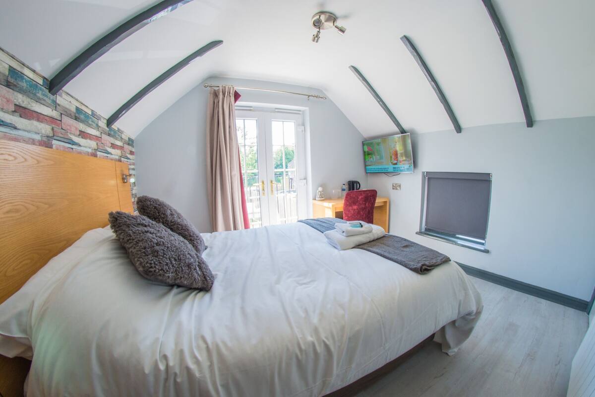 A bedroom with a sloped ceiling, a bed with pillows, a TV, a window, and a wooden wardrobe.