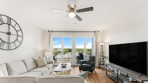 A modern living room with a large sofa, a television, and a ceiling fan. - Lakefront resort condo w/ pools & daily tickets (Panama City Beach)